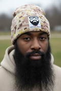 Two camouflage knit beanie hats in shades of beige, brown, and muted yellow, laid flat on artificial green grass. Each beanie has a white square patch with a black beard and sunglasses graphic and the text 'BEARD GANG CHICAGO'.